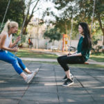 two young women sit on swing set and talk while laughing